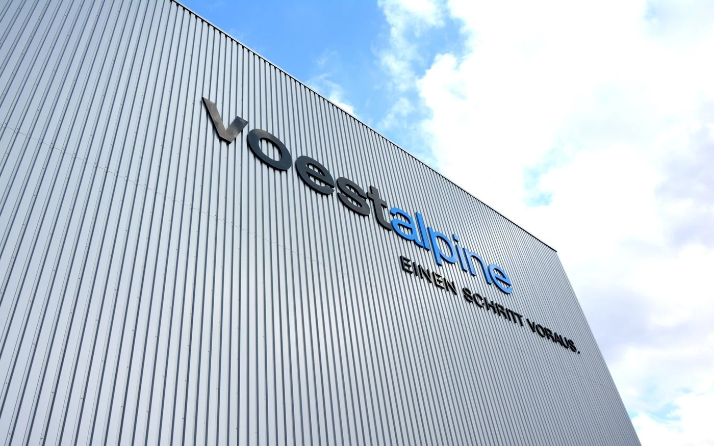 Schrägansicht eines großen Industriegebäudes mit vertikaler Metallverkleidung, entworfen in Zusammenarbeit mit SCHATZ Architektur. An der Seite sind der Schriftzug „voestalpine“ und der Slogan „EINEN SCHRITT VORAUS“ zu sehen. Der Himmel ist teilweise bewölkt.