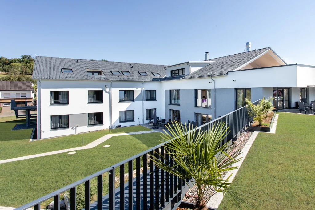 Ein modernes, weißes Gebäude mit großen Fenstern, einem grauen Dach und einem üppig grünen Rasen, gesäumt von schwarzen Geländern und kleinen Palmen, unter einem strahlend blauen Himmel – ein perfektes Beispiel für zeitgenössische Projektentwicklung und Architektur in Schorndorf.