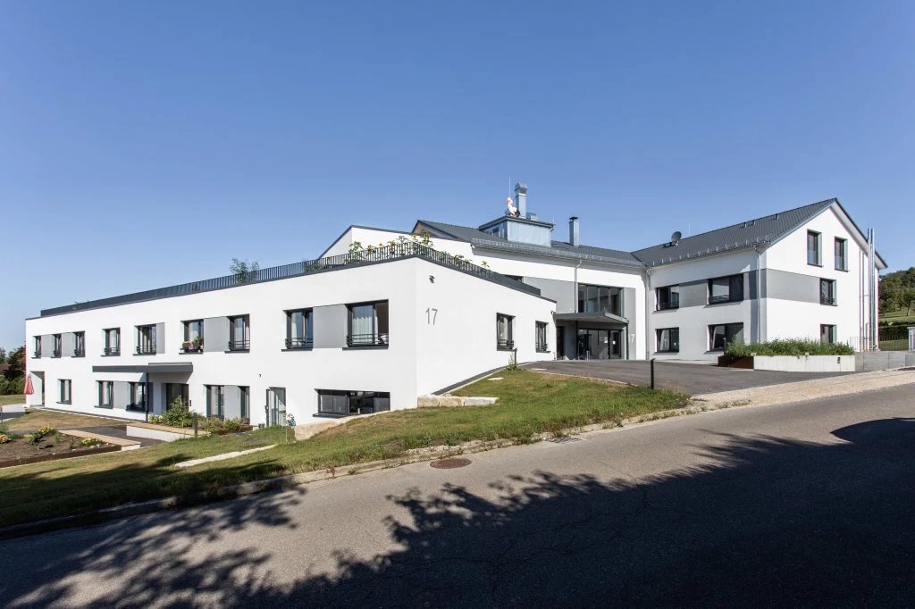 Pflege Ein modernes, weißes, zweistöckiges Gebäude mit vielen Fenstern und einem schrägen Dach, das auf einem grasbewachsenen Hang unter strahlend blauem Himmel steht, ist ein Paradebeispiel für innovative Projektentwicklung in Schorndorf.