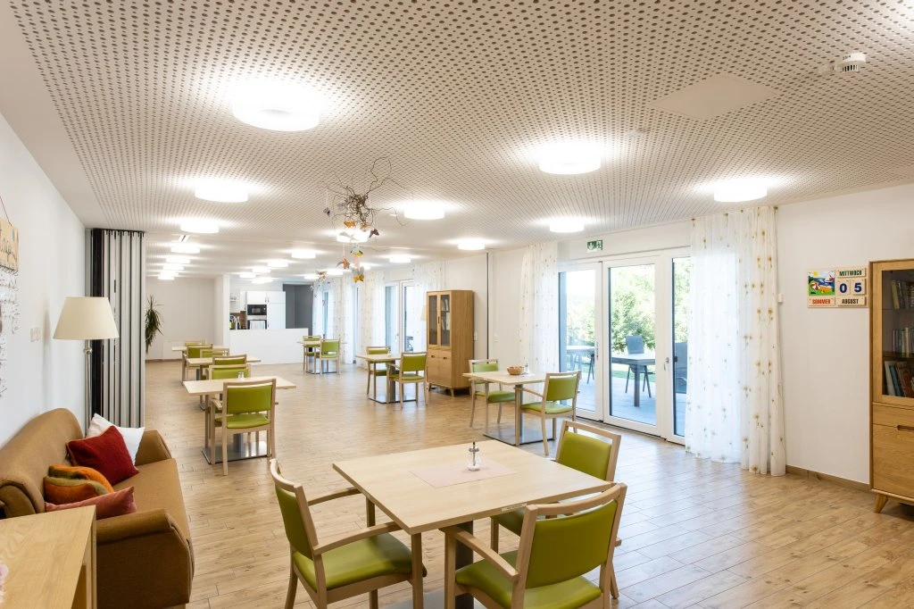 Ein heller, geräumiger Raum mit Holzböden, grünen Stühlen um quadratische Tische, einem Sofa, großen Fenstern mit weißen Vorhängen und modernen Deckenleuchten – ein einladender Raum, der von SCHATZ Architektur meisterhaft gestaltet wurde. Im Hintergrund sind einige Regale und Dekorationsgegenstände zu sehen, die den durchdachten Ansatz des Büros bei der Generalplanung über alle Leistungsphasen hinweg widerspiegeln.