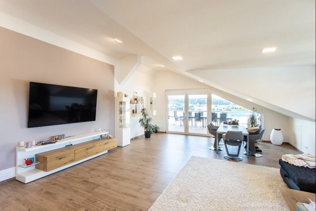 Ein modernes Wohnzimmer in Schorndorf mit einem an der Wand montierten Fernseher, Holzböden, einem Essbereich, großen Fenstern, die auf einen Balkon führen, hellen Wänden und zeitgemäßen Möbeln unter einer schrägen Decke – ein einladender Raum, der die durchdachte Projektentwicklung über alle Leistungsphasen hinweg perfekt zur Geltung bringt.