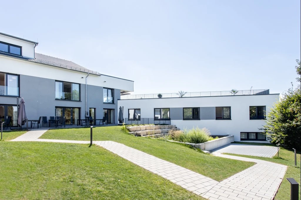 Ein modernes zweistöckiges Gebäude mit großen Fenstern, einem Terrassenbereich mit Tischen und Stühlen, umgeben von grünem Rasen, gepflasterten Wegen, einer gepflegten Außenanlage und dem blauen Himmel im Hintergrund – ein hervorragendes Beispiel für innovativen Entwurf und durchdachte Projektentwicklung in Schorndorf.