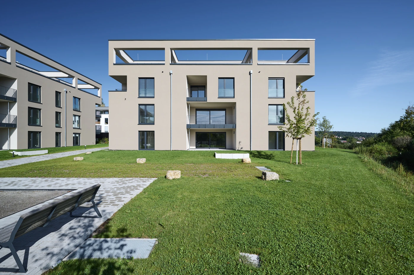 Projekte In Schorndorf steht ein modernes dreistöckiges Mehrfamilienhaus mit großen Fenstern und Flachdächern, umgeben von einem gepflegten Rasen, Bänken und jungen Bäumen unter strahlend blauem Himmel – ein beeindruckendes Ergebnis einer durchdachten Projektentwicklung über alle Leistungsphasen hinweg.