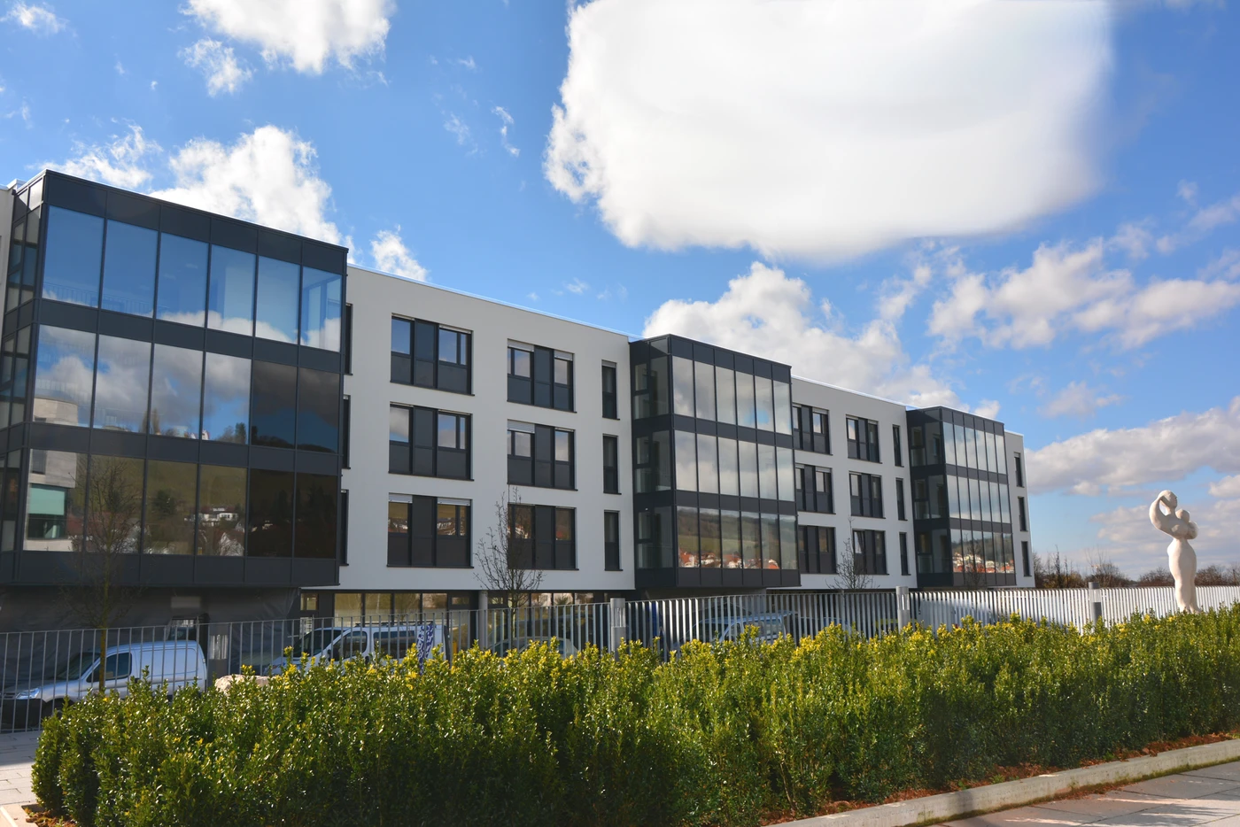Ein modernes Bürogebäude aus Weiß und Glas mit mehreren Stockwerken, großen Fenstern, einem Metallzaun, grünen Sträuchern davor, geparkten Autos und einem bewölkten blauen Himmel im Hintergrund – ein ideales Beispiel für zeitgemäße Generalplanung und durchdachte Projektentwicklung.
