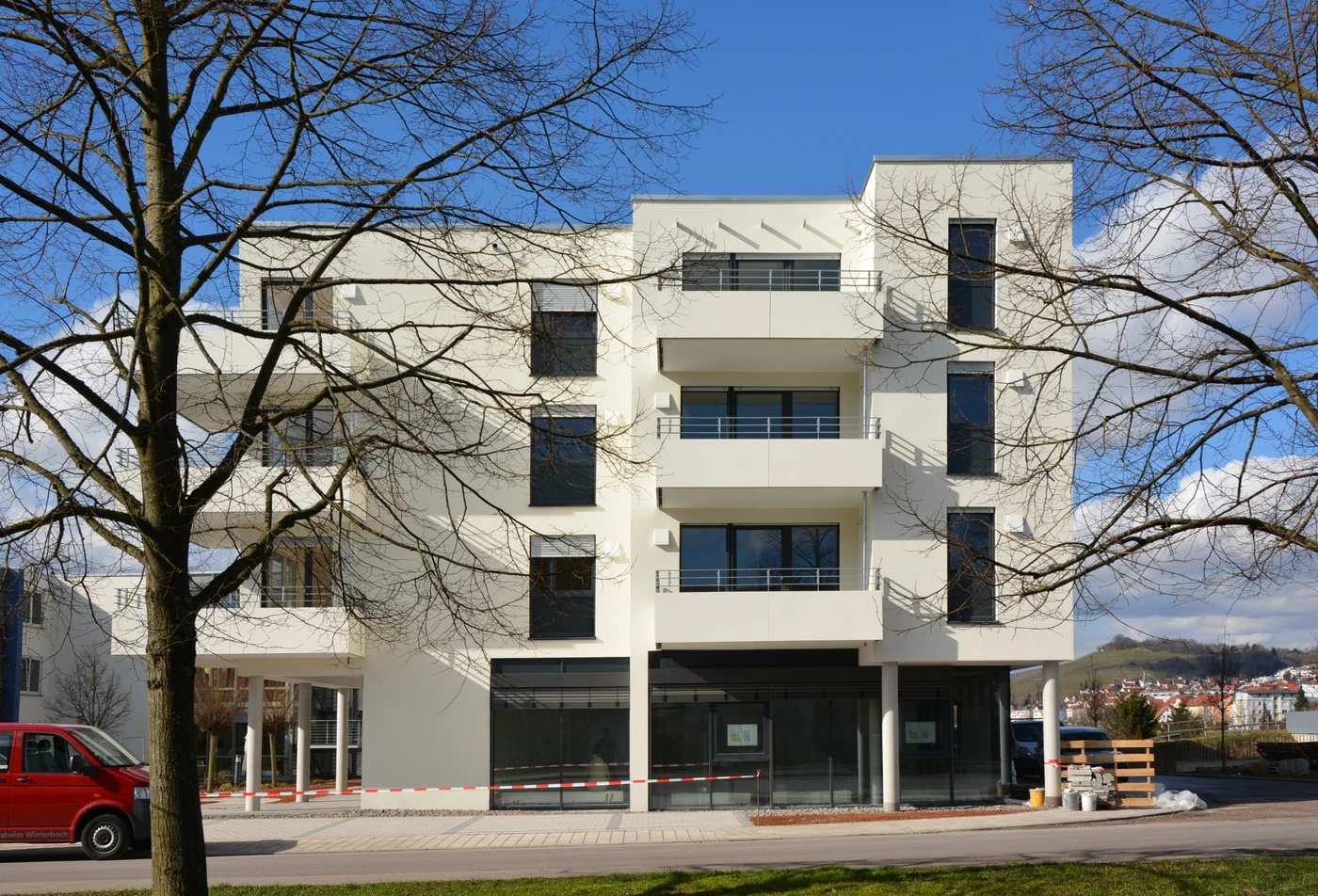 Modernes weißes Mehrfamilienhaus mit Balkonen und großen Fenstern, das teilweise im Schatten blattloser Bäume liegt. Links steht ein roter Transporter, rechts liegen Baumaterialien, was auf laufende Bauarbeiten hindeutet. Dieses zeitgenössische Gebäude ist ein Beispiel für die durchdachte Projektentwicklung von SCHATZ Architektur. Blauer Himmel mit einigen Wolken.