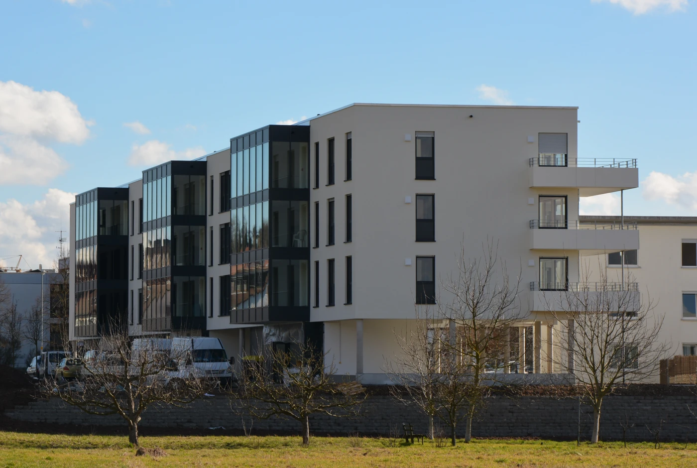 Ein modernes Mehrfamilienhaus mit großen Glasfenstern und Balkonen, entworfen von SCHATZ Architektur, steht umgeben von kahlen Bäumen und einer Rasenfläche unter einem blauen Himmel mit einigen Wolken.