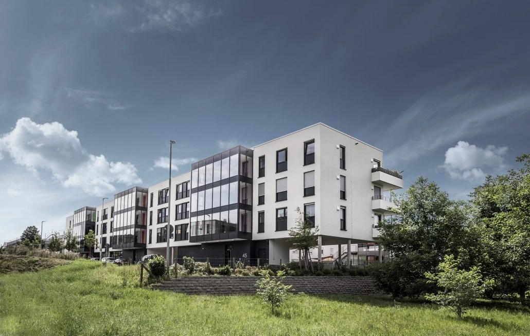 Pflege Ein modernes, mehrstöckiges Wohnhaus mit großen Fenstern und Balkonen zeugt von innovativem architektonischem Entwurf und liegt, umgeben von grünem Rasen und Bäumen unter einem teilweise bewölkten Himmel – ein hervorragendes Beispiel für zeitgenössische Projektentwicklung in Schorndorf.