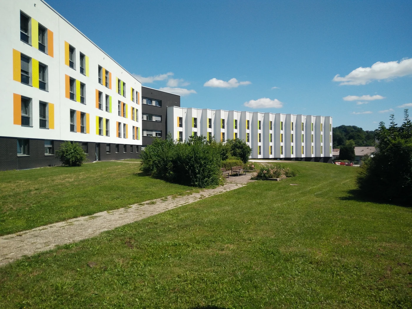 Pflege Ein modernes, mehrstöckiges Gebäude mit weißen und gelben Fassadenplatten, umgeben von grünem Rasen, Sträuchern und einem Steinweg unter einem sonnigen blauen Himmel mit wenigen Wolken – ein eindrucksvolles Beispiel für Generalplanung über alle Leistungsphasen hinweg im Herzen von Schorndorf.