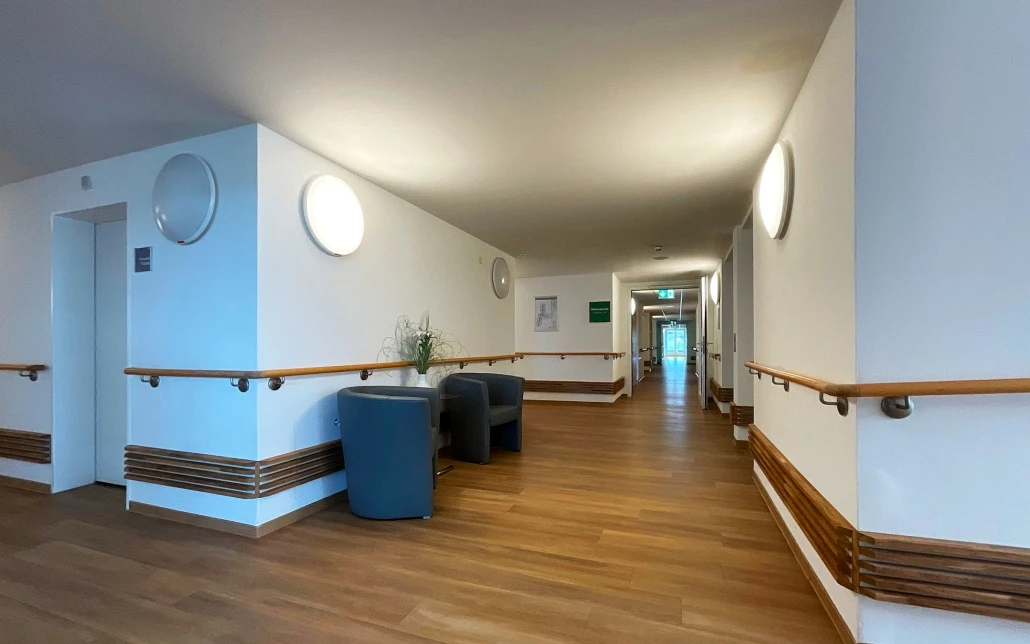 Projekte Ein heller, klar gestalteter Flur, entworfen von SCHATZ Architektur, besticht durch Holzböden, weiße Wände, Handläufe sowie zwei dunkle Stühle mit einem kleinen Tisch und einer Pflanze. Entlang des Flurs sind Türen und runde Wandleuchten zu sehen, die die durchdachte Projektentwicklung und die Liebe zum Detail in der Generalplanung widerspiegeln.