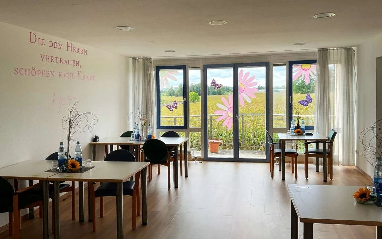 Pflege Ein heller Raum mit großen Fenstern und Blick auf die Felder, geschmückt mit Blumen- und Schmetterlingsaufklebern. Vier Schreibtische mit Flaschen und Sonnenblumen stehen in großem Abstand zueinander; an der Wand ist ein deutscher Text zu sehen, der den durchdachten Entwurf von SCHATZ Architektur im Rahmen der Generalplanung widerspiegelt.