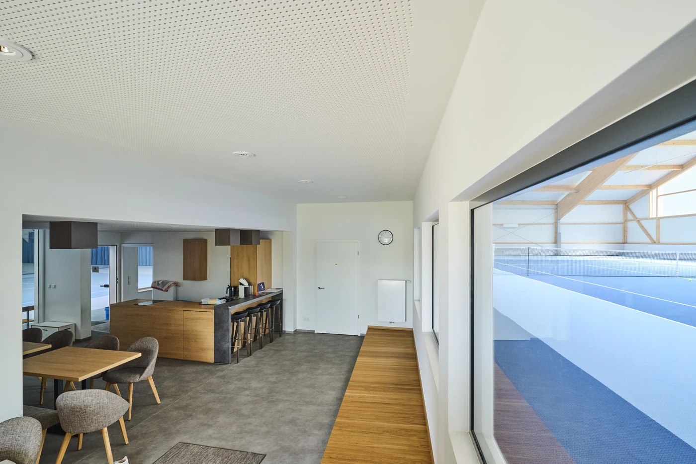 Projekte Modernes, helles Zimmer mit Küche, Essbereich, Holzmöbeln und großen Fenstern mit Blick auf eine Tennishalle in Schorndorf. Der Raum besticht durch eine minimalistische Einrichtung und viel Tageslicht und zeugt von einem durchdachten Entwurf und viel Liebe zum Detail bei der Gesamtplanung.