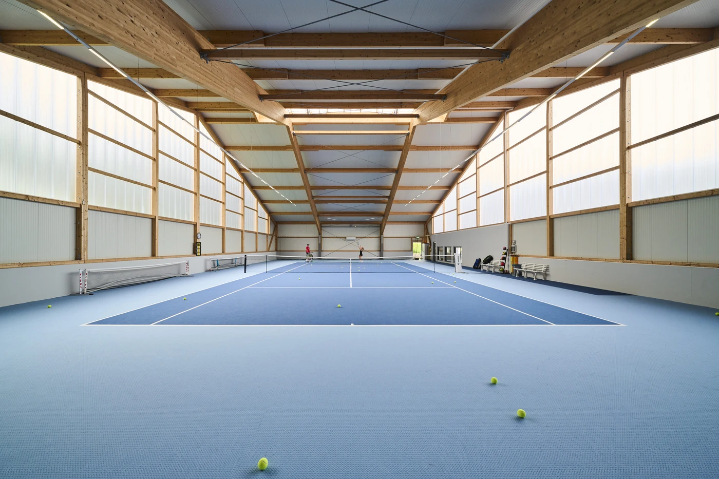 Projekte Ein Tennishalle mit blauem Belag, mehreren Tennisbällen auf dem Boden und Menschen im Hintergrund in der Nähe von Bänken unter einer hohen Decke mit Holzbalken zeigt einen beeindruckenden Entwurf, der in Schorndorf im Rahmen verschiedener Leistungsphasen realisiert wurde.
