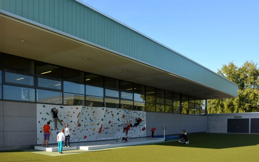 Kinder spielen und klettern an einer Kletterwand im Freien, die an einem modernen Gebäude angebracht ist – ein beeindruckendes Ergebnis durchdachter Planung und sorgfältiger Projektentwicklung –, während die Erwachsenen in der Nähe auf einer Rasenfläche unter strahlend blauem Himmel ein Auge auf sie haben.