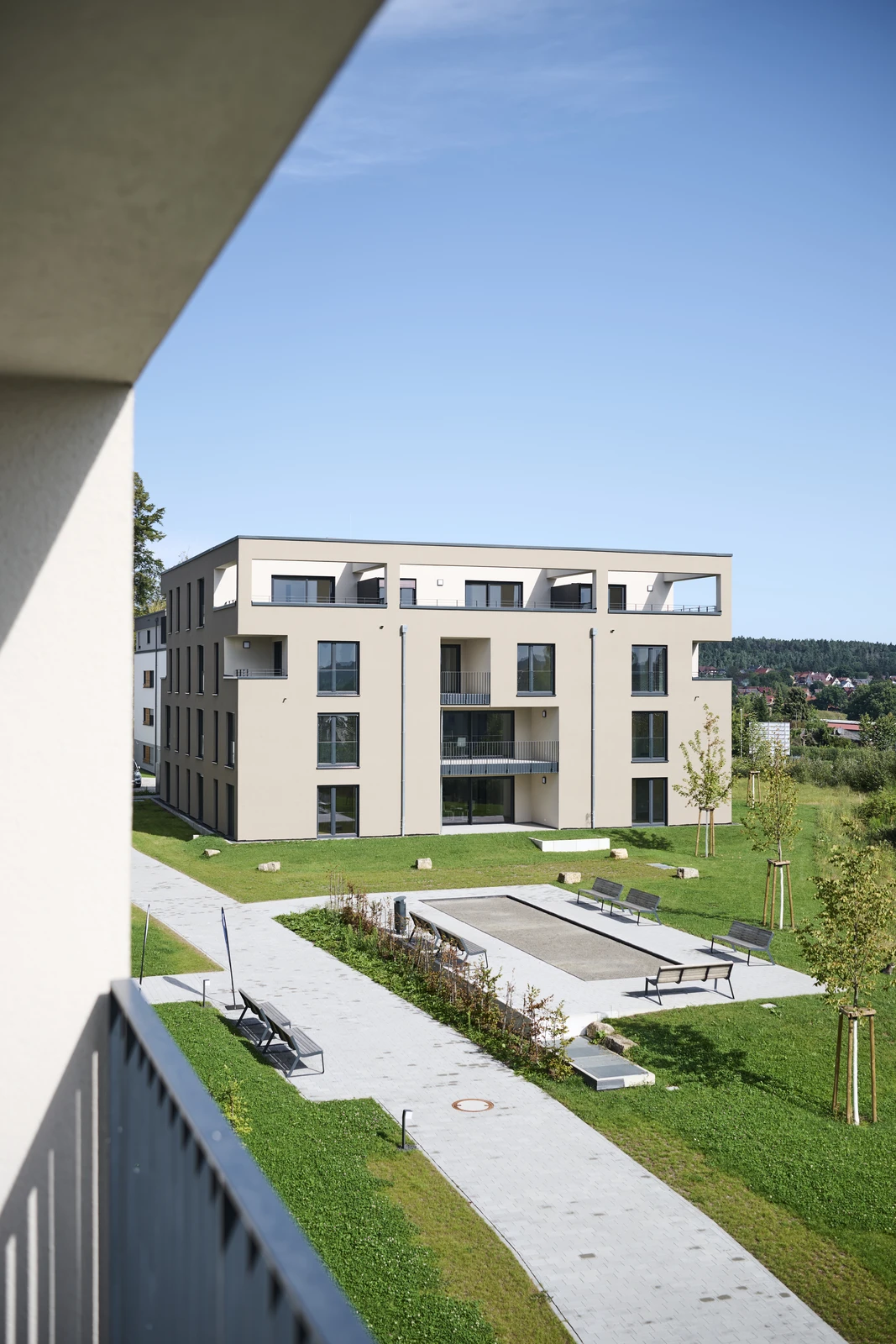 Ein modernes, dreistöckiges, beiges Wohnhaus mit Balkonen, großen Fenstern und einem begrünten Innenhof mit Bänken, Bäumen und grünen Rasenflächen unter strahlend blauem Himmel zeugt von durchdachtem Entwurf und sorgfältiger Gesamtplanung in allen Leistungsphasen.
