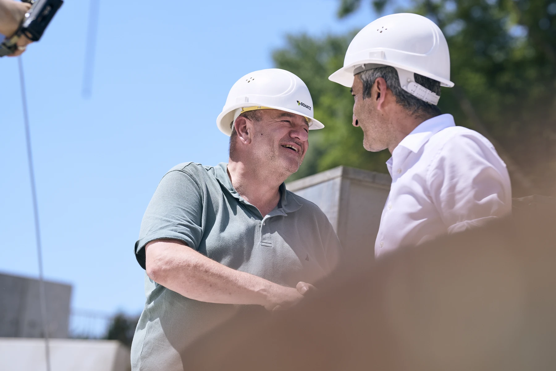 Startseite Zwei Männer mit weißen Bauhelmen geben sich an einem sonnigen Tag im Freien die Hand und lächeln einander an. Der eine trägt ein grünes Poloshirt, der andere ein weißes Hemd; beide vertreten SCHATZ Architektur, während sie einen Entwurf für die kommenden Leistungsphasen besprechen. Im Hintergrund sind Bäume und der blaue Himmel zu sehen.