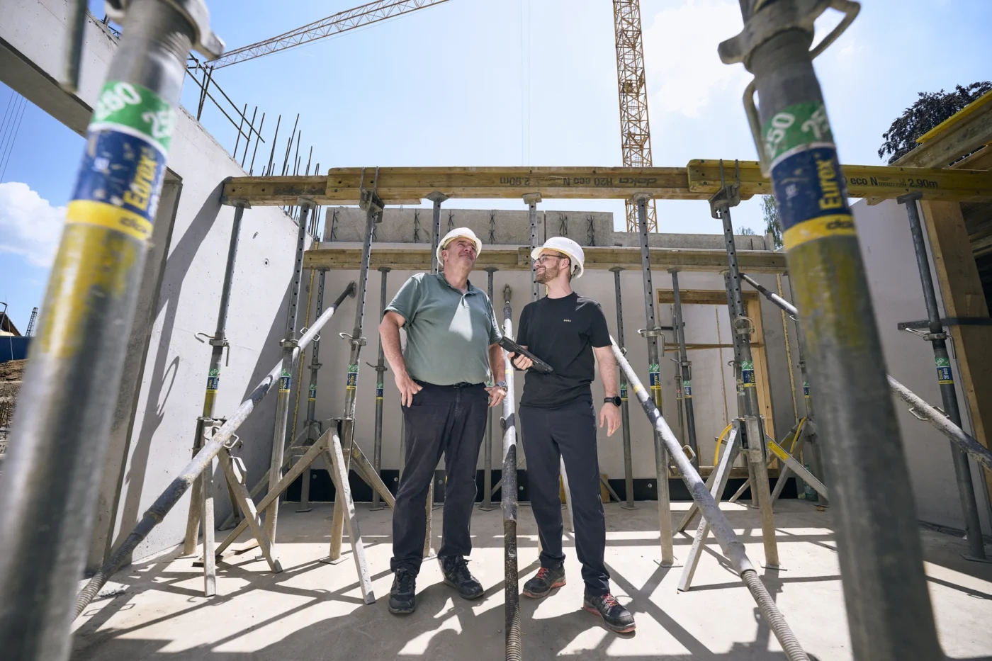 Team Zwei Personen mit Schutzhelmen unterhalten sich auf einer Baustelle, umgeben von Metallstützbalken; im Hintergrund sind unter blauem Himmel das Rohbaugerüst und ein Kran zu sehen – eine typische Szene während der von SCHATZ Architektur geleiteten Entwurfs- und Projektentwicklungsphasen.