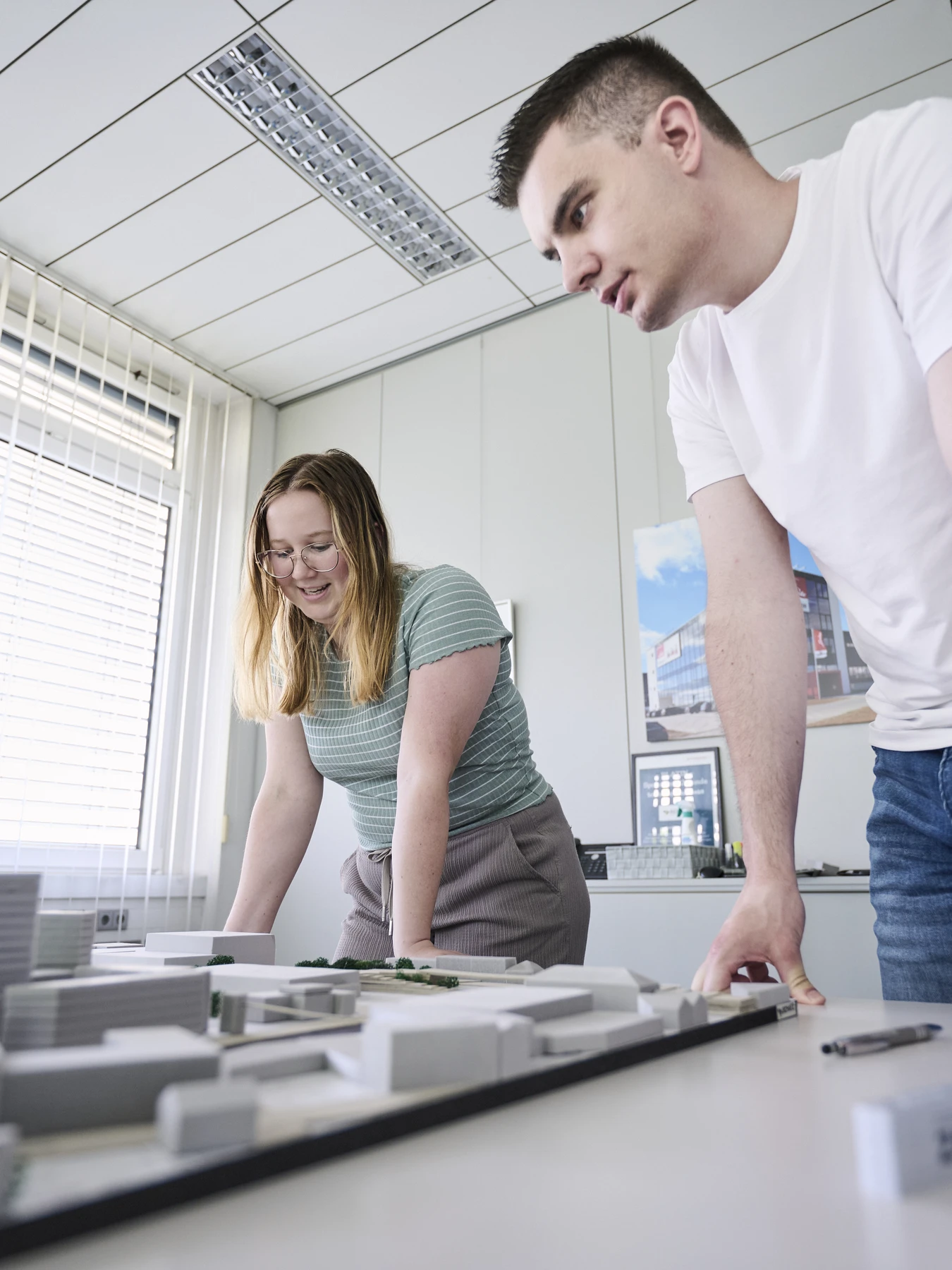 Startseite Zwei Personen beugen sich über einen Tisch und betrachten ein Architekturmodell von Gebäuden in einem hellen Büro in Schorndorf mit großen Fenstern und gerahmten Bildern an den Wänden; sie arbeiten gemeinsam an dem nächsten Entwurf in den frühen Leistungsphasen.