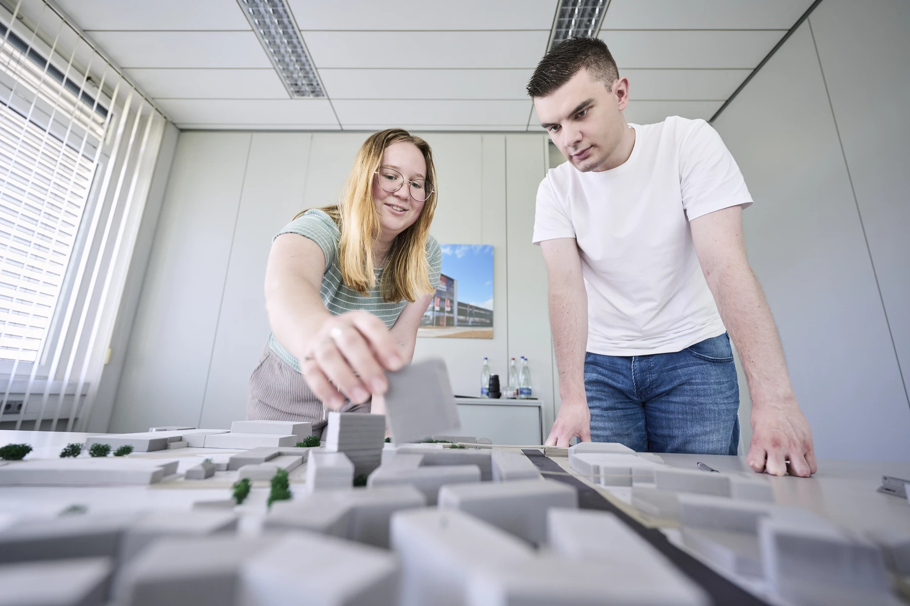 Projektanfrage Zwei Personen begutachten und passen Teile eines grauen Architekturmodells einer Stadt an – in einem hellen Büro mit großen Fenstern und Deckenleuchten. So wird der Geist der Zusammenarbeit zwischen Entwurf und Projektentwicklung eingefangen, während die beiden ihre Ideen im Rahmen der wichtigsten Leistungsphasen verfeinern.