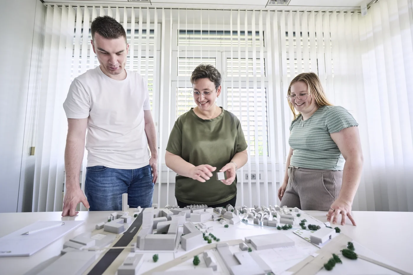 Was uns ausmacht Drei Personen stehen um einen Tisch herum, betrachten ein weißes Architekturmodell eines Stadtviertels und besprechen es in einem hellen Raum mit Vertikaljalousien. Die Gruppe begutachtet den innovativen Entwurf von SCHATZ Architektur und erörtert Ideen für das bevorstehende Projekt in Schorndorf.