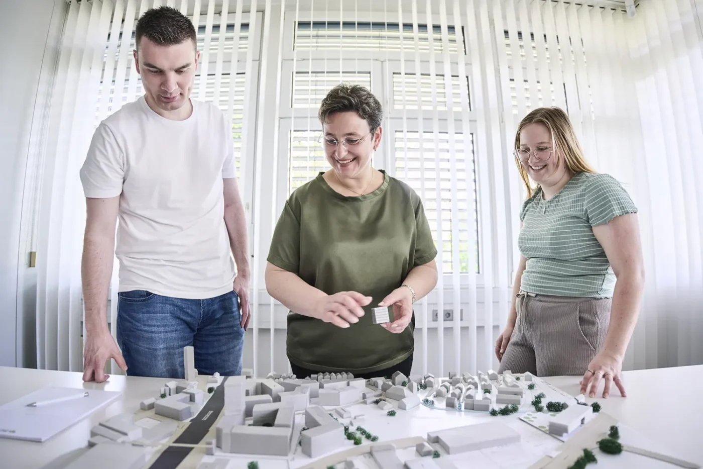 Was uns ausmacht Drei Personen stehen um einen Tisch herum, betrachten ein weißes Architekturmodell eines Stadtviertels und besprechen es in einem hellen Raum mit Vertikaljalousien. Die Gruppe begutachtet den innovativen Entwurf von SCHATZ Architektur und erörtert Ideen für das bevorstehende Projekt in Schorndorf.