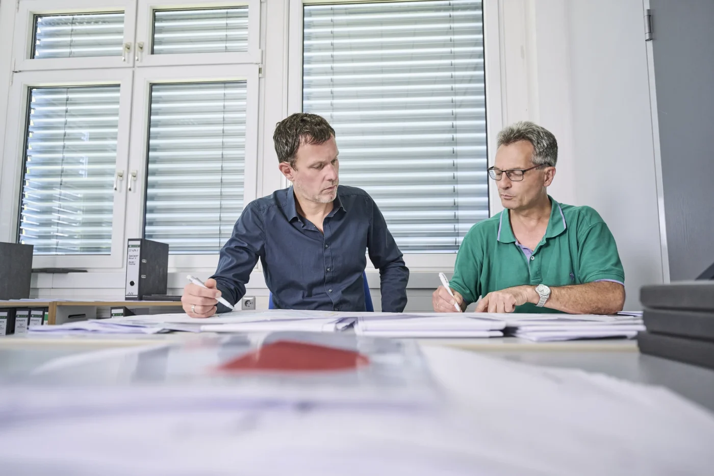 Was uns ausmacht Zwei Männer sitzen an einem mit Unterlagen bedeckten Tisch, besprechen die Generalplanung und machen sich gemeinsam Notizen in einem hellen Büro mit großen Fenstern und Jalousien im Hintergrund; wahrscheinlich gehen sie dabei verschiedene Leistungsphasen für ein Projekt in Schorndorf durch.