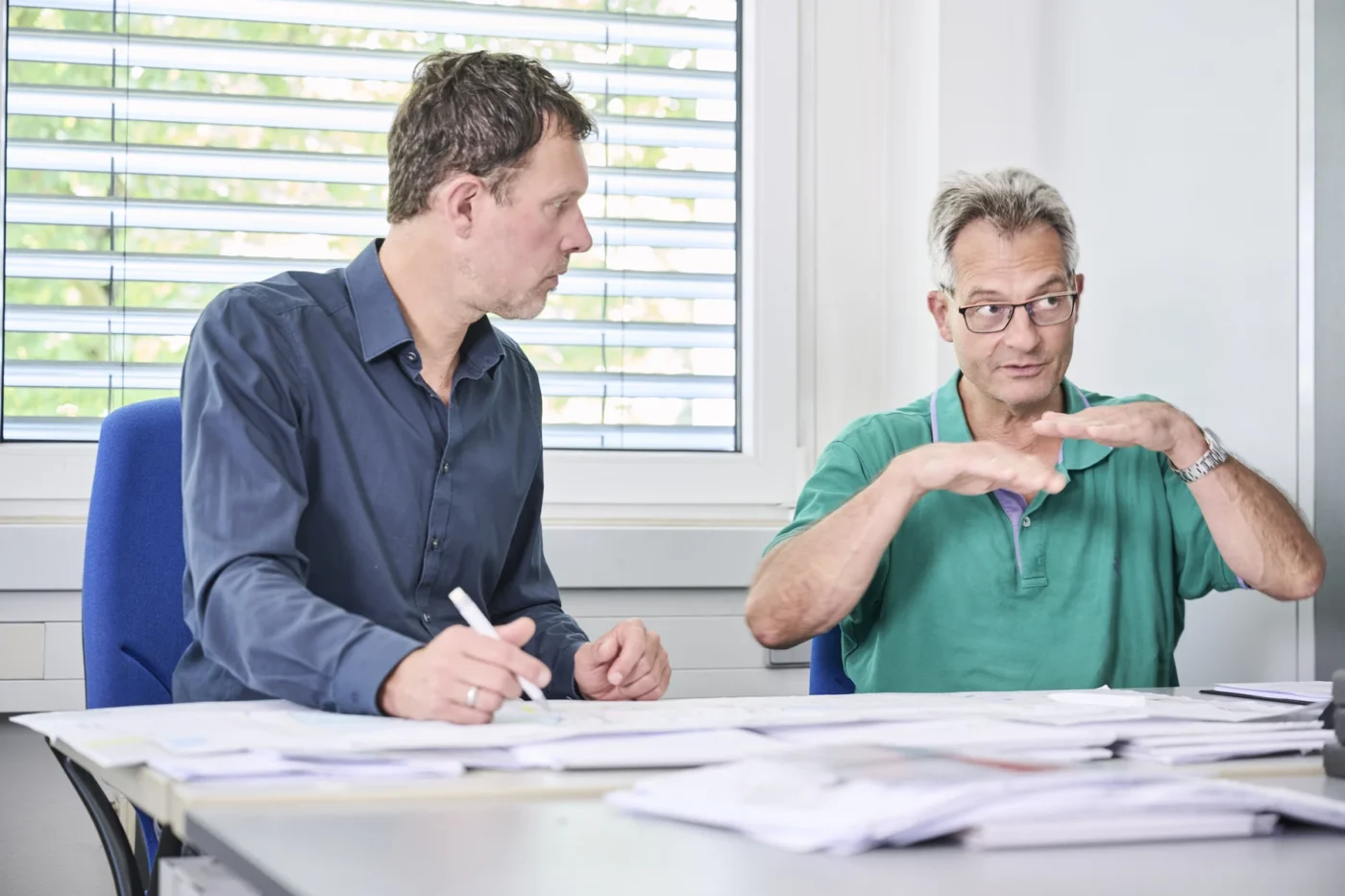 Sport Zwei Männer sitzen an einem mit Papieren bedeckten Tisch; der eine erklärt etwas mit Handgesten, während der andere zuhört und sich Notizen macht. Sie scheinen in einem hellen Büro, das sich möglicherweise in Schorndorf befindet, ein konzentriertes Gespräch über Projektentwicklung und Leistungsphasen zu führen.