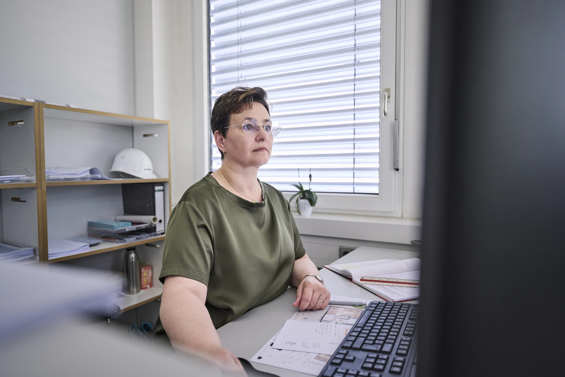 Pflege Eine Frau mit kurzen Haaren und Brille sitzt an einem Schreibtisch in einem Büro und blickt auf einen Computerbildschirm. Papiere, eine Tastatur und Regale mit Unterlagen im Hintergrund lassen vermuten, dass sie sich im Rahmen ihrer Aufgaben mit Projektentwicklung oder der Leitung verschiedener Leistungsphasen beschäftigt.