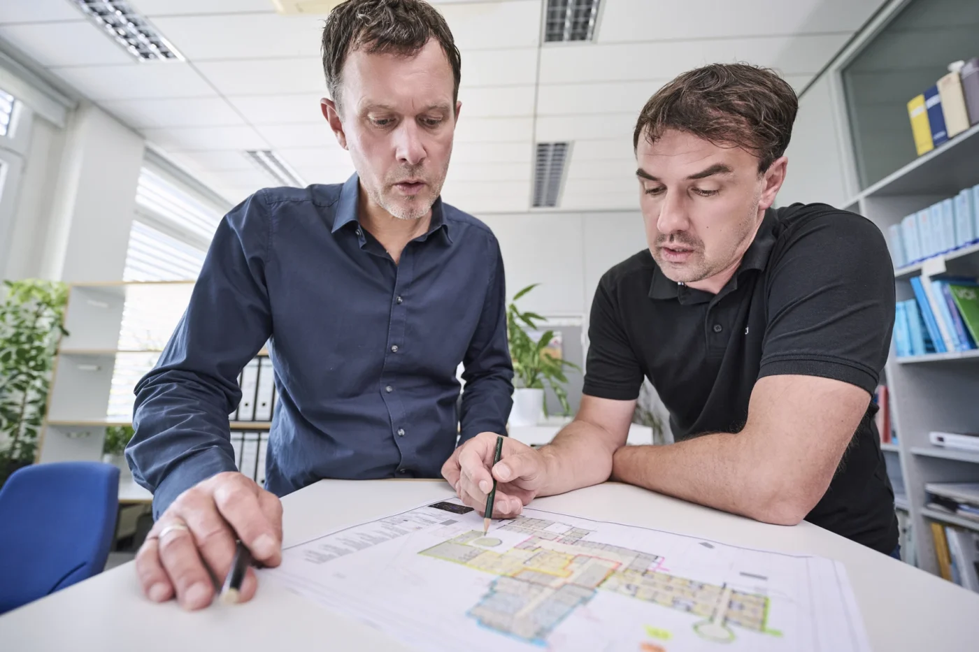 Industrie & Gewerbe Zwei Männer besprechen an einem Schreibtisch in einem modernen Büro einen Grundriss und sehen sich einen Entwurf von SCHATZ Architektur an. Beide zeigen auf die Bauzeichnung und konzentrieren sich auf die Details – vielleicht stellen sie sich dabei ihr nächstes Projekt in Schorndorf vor. Im Hintergrund sind Regale mit Büchern und Pflanzen zu sehen.