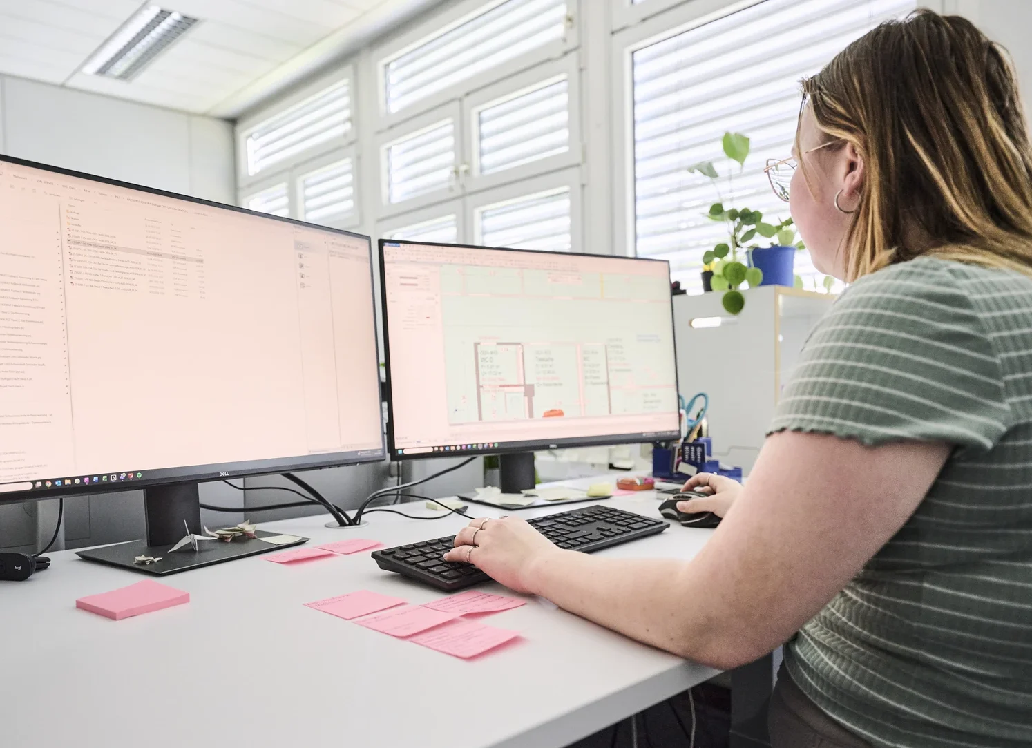 Pflege Eine Person mit langen Haaren und Brille sitzt an einem Schreibtisch mit zwei großen Computermonitoren, umgeben von rosa Haftnotizen und Büromaterial, in einem hell beleuchteten Raum mit großen Fenstern und konzentriert sich intensiv auf die Projektentwicklung für SCHATZ Architektur in Schorndorf.