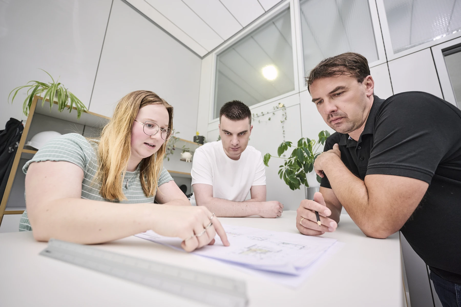 Projektanfrage Drei Personen sitzen an einem Tisch in einem modernen Büro und gehen gemeinsam Unterlagen zum Thema Projektentwicklung durch. Eine Frau zeigt auf die Unterlagen, während zwei Männer aufmerksam zuschauen; sie arbeiten als Teil des Teams von SCHATZ Architektur zusammen. Im Hintergrund sind Pflanzen und Regale zu sehen.