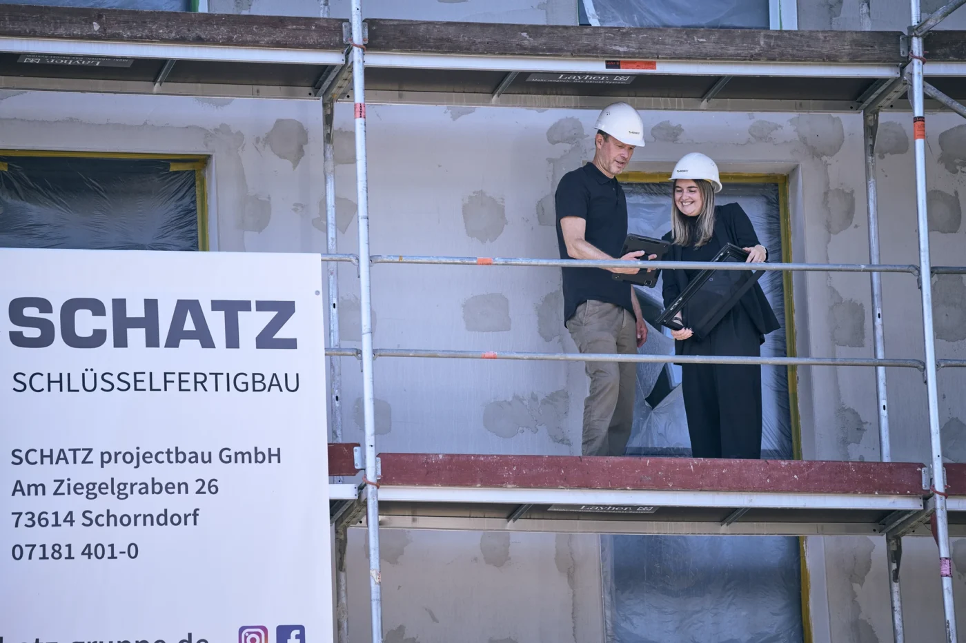 Über uns Zwei Personen mit Schutzhelmen stehen auf einem Gerüst auf einer Baustelle in Schorndorf und schauen auf ein Klemmbrett. Ein großes Schild in der Nähe zeigt den Firmennamen, die Adresse und die Kontaktdaten von SCHATZ Architektur an, was darauf hindeutet, dass es sich hier um die Baustelle ihres neuesten Entwurfs handelt.