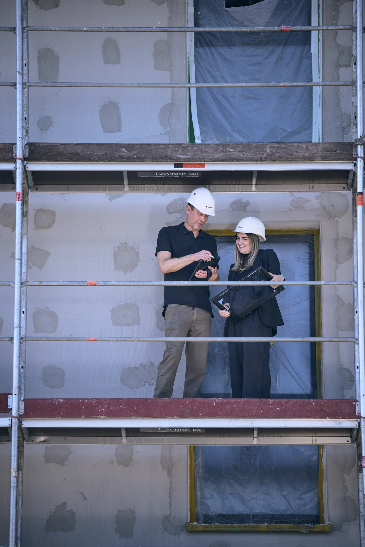 Zwei Personen mit Schutzhelmen stehen auf einem Gerüst auf einer Baustelle und schauen gemeinsam auf ein Tablet, während sie den Fortschritt eines Projekts von SCHATZ Architektur Projektentwicklung in Schorndorf besprechen. Das Gebäude hinter ihnen hat unfertige Wände und mit Plastikfolie abgedeckte Fenster.
