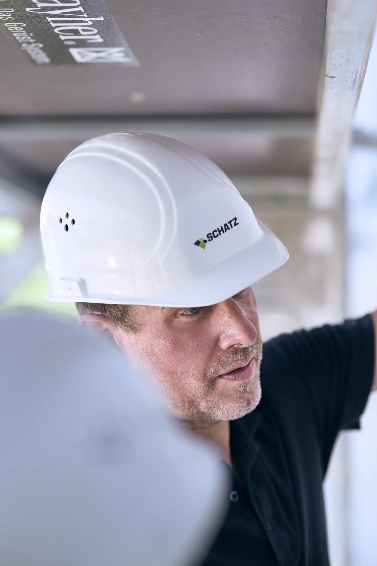 Sport Ein Mann mit einem weißen Schutzhelm, auf dem „SCHATZ“ steht, blickt nach oben, während er auf einer Baustelle arbeitet. Als Vertreter von SCHATZ Architektur wirkt er konzentriert und wird teilweise von Geräten im Vordergrund verdeckt.