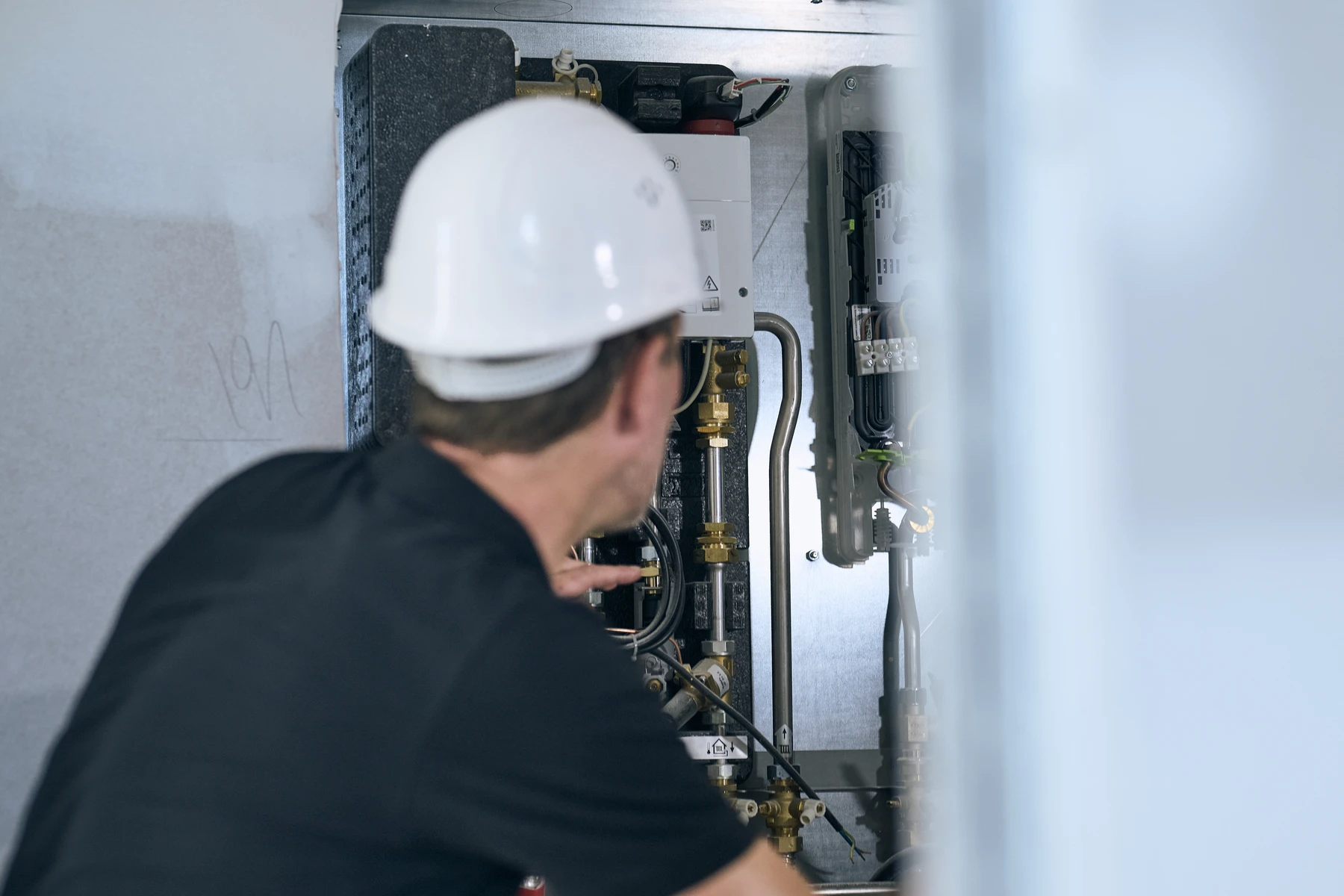Startseite Eine Person mit einem weißen Schutzhelm und einem schwarzen Hemd inspiziert oder arbeitet an Industrieanlagen mit Rohren und Ventilen in einem Metallschrank, wahrscheinlich im Rahmen einer Projektentwicklung oder Generalplanung in Schorndorf. Das Gesicht der Person ist nicht zu sehen, der Fokus liegt auf den Maschinen.