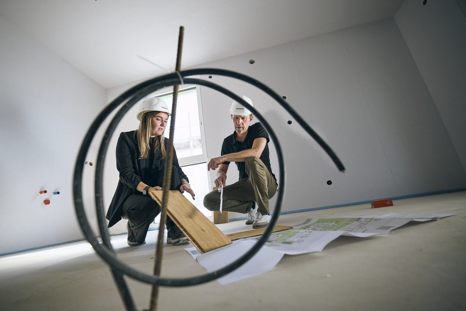 Was uns ausmacht Zwei Personen mit Schutzhelmen begutachten Holzbretter und Baupläne auf dem Boden einer Baustelle – aufgenommen durch ein rundes Metallrohr –, während sie unter der fachkundigen Anleitung des Generalplanungsteams von SCHATZ Architektur die aktuellen Leistungsphasen besprechen.