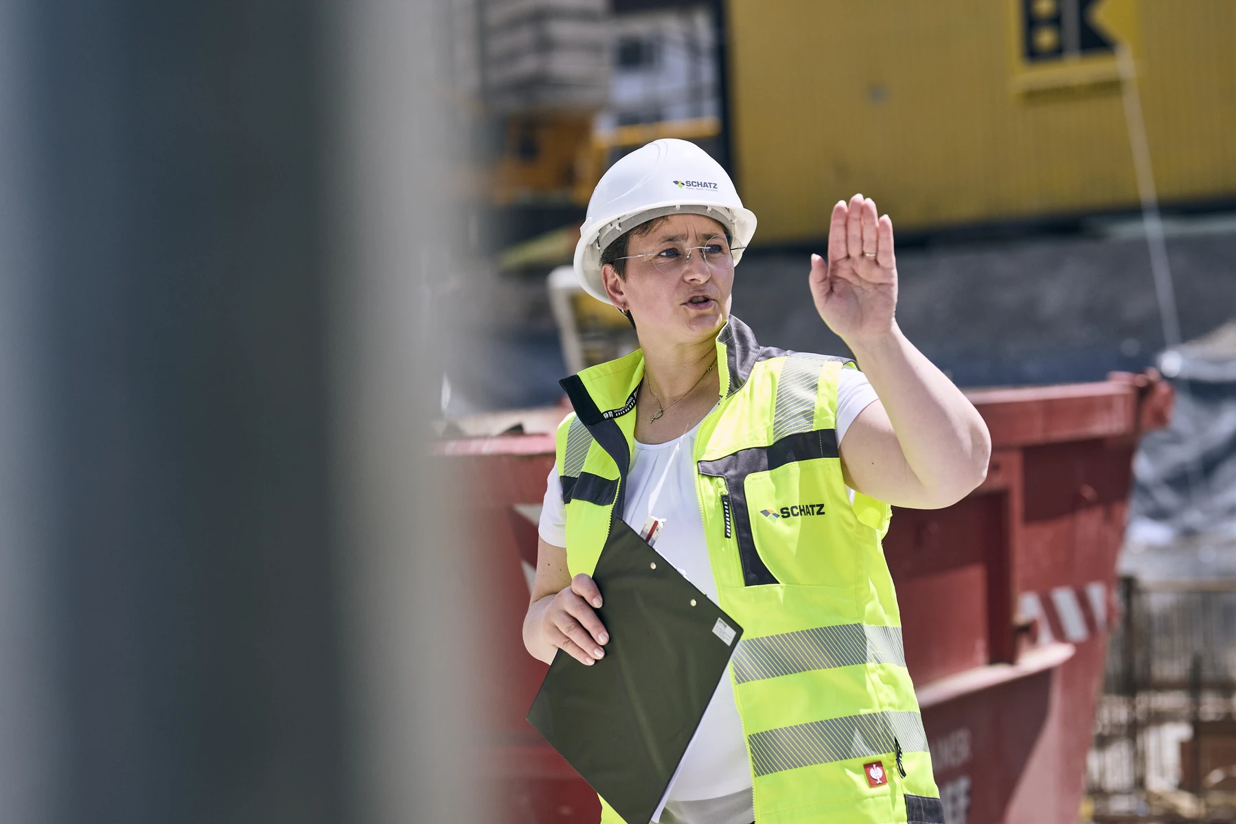 Bildung Ein Bauarbeiter in einer gelben Sicherheitsweste und einem weißen Schutzhelm gestikuliert mit einer Hand, während er ein Klemmbrett in der Hand hält. Er steht auf einer Baustelle von SCHATZ Architektur Projektentwicklung in Schorndorf, im Hintergrund sind Baugeräte und Baumaterialien zu sehen.