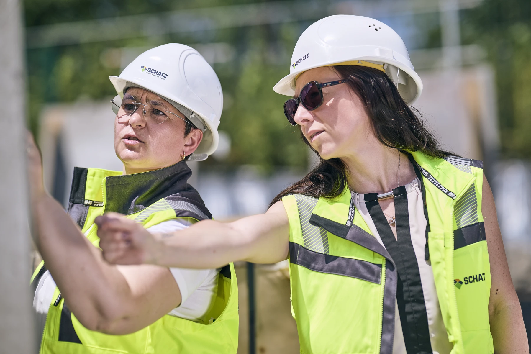 Neubau Pflegeheim in Urbach Zwei Frauen mit Schutzhelmen und Sicherheitswesten besprechen auf einer Baustelle die Pläne und gehen dabei den Entwurf in einer der frühen Leistungsphasen durch. Die eine zeigt nach vorne, während die andere aufmerksam zusieht. Beide sind konzentriert und engagiert bei der Arbeit und zeigen damit ihr Engagement für eine präzise Generalplanung.