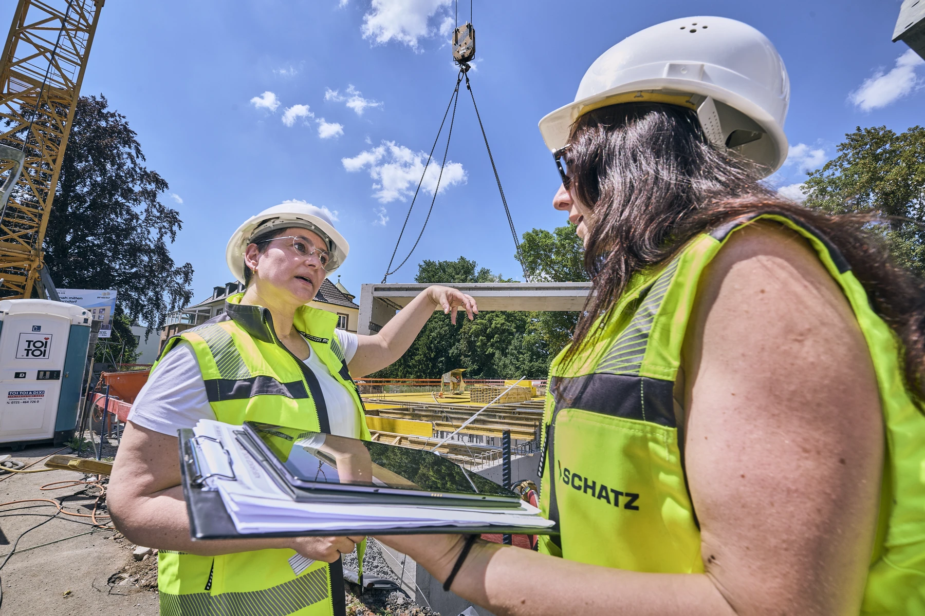 Industrie & Gewerbe Zwei Frauen in Sicherheitswesten und Helmen unterhalten sich auf einer Baustelle in Schorndorf. Die eine hält ein Tablet und einen Schreibblock in der Hand – unverzichtbare Hilfsmittel für die Abwicklung verschiedener Leistungsphasen –, während die andere auf einen Kran deutet, der im Hintergrund einen großen Träger hebt. Der blaue Himmel und die Bäume bilden eine inspirierende Kulisse für ihre Entwurfsgespräche.