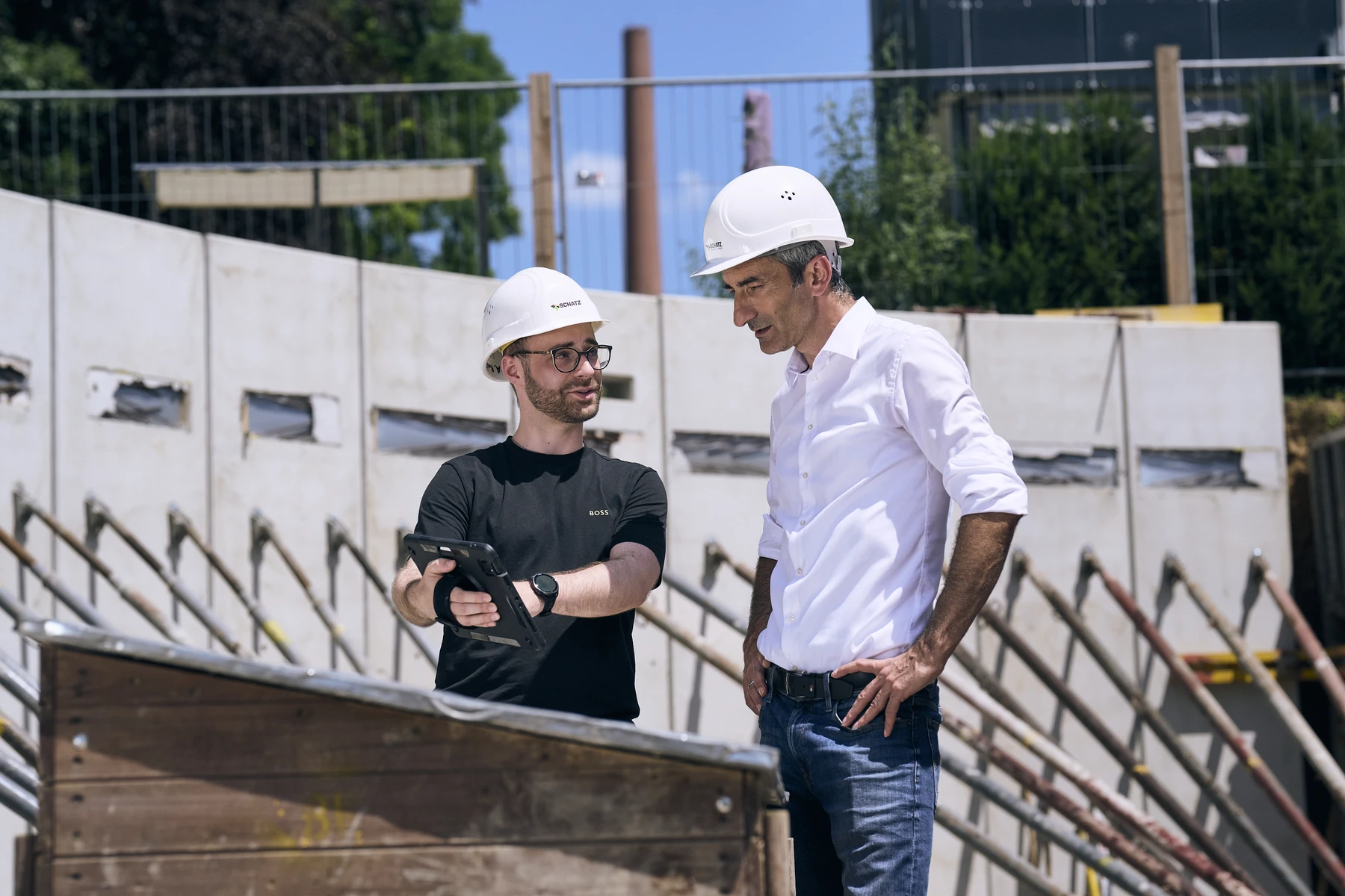 Was uns ausmacht Zwei Männer mit weißen Schutzhelmen stehen auf einer Baustelle in Schorndorf. Der eine hält ein Tablet in der Hand und gestikuliert, während er mit dem anderen über den aktuellen Entwurf spricht. Beide tragen Hemden und sind in ein Gespräch vertieft; im Hintergrund sind Baumaterialien zu sehen.