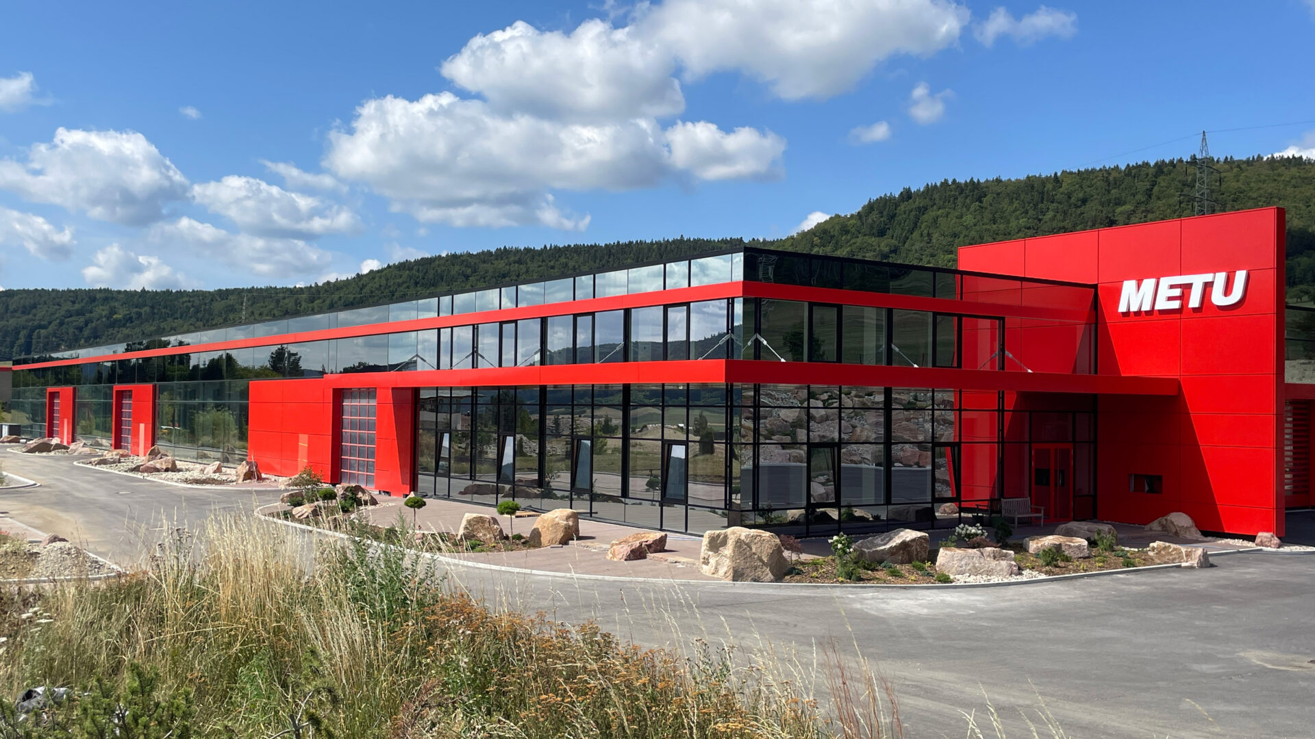 Architektur Ein modernes Gebäude mit einer Fassade aus rotem Material und Glas, auf dem „METU“ steht, befindet sich neben einer Straße mit Grünanlagen und Felsen, vor dem Hintergrund von Bäumen und einem blauen Himmel mit Wolken.