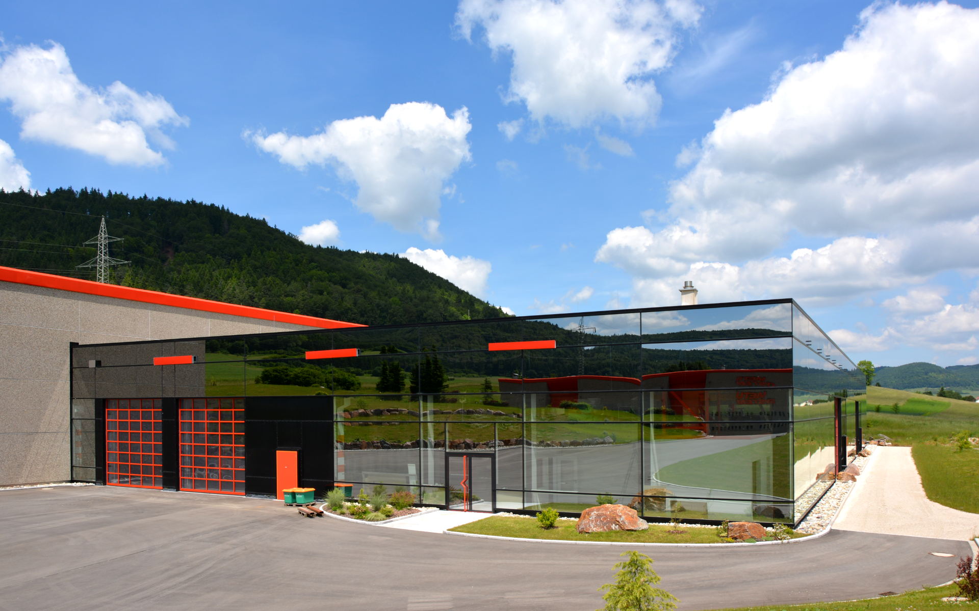 Ein modernes Gebäude mit großen Spiegelglaswänden spiegelt die grüne Landschaft und den blauen Himmel wider. Das Gebäude weist rote Akzente auf und ist unter teilweise bewölktem Himmel von Hügeln, Wiesen und Bäumen umgeben.