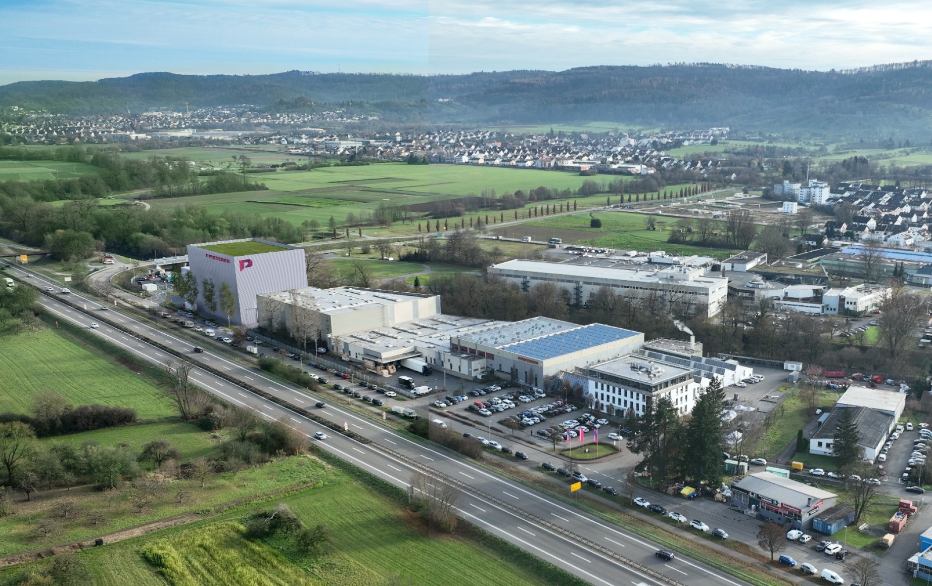 Ein großer Industriekomplex mit weißen Gebäuden und einem hohen, mit rosa Akzenten versehenen Bauwerk liegt an einer vielbefahrenen Autobahn, umgeben von grünen Feldern und Hügeln unter einem teilweise bewölkten Himmel.