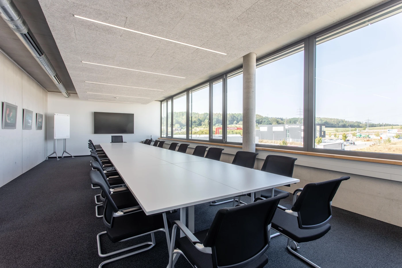 Ein moderner Konferenzraum von SCHATZ Architektur in Schorndorf verfügt über einen langen Tisch, schwarze Stühle, große Fenster mit Blick ins Freie, ein Whiteboard und einen an der Wand montierten Fernseher. Der helle und minimalistische Raum spiegelt eine durchdachte Gestaltung über alle Leistungsphasen hinweg wider.