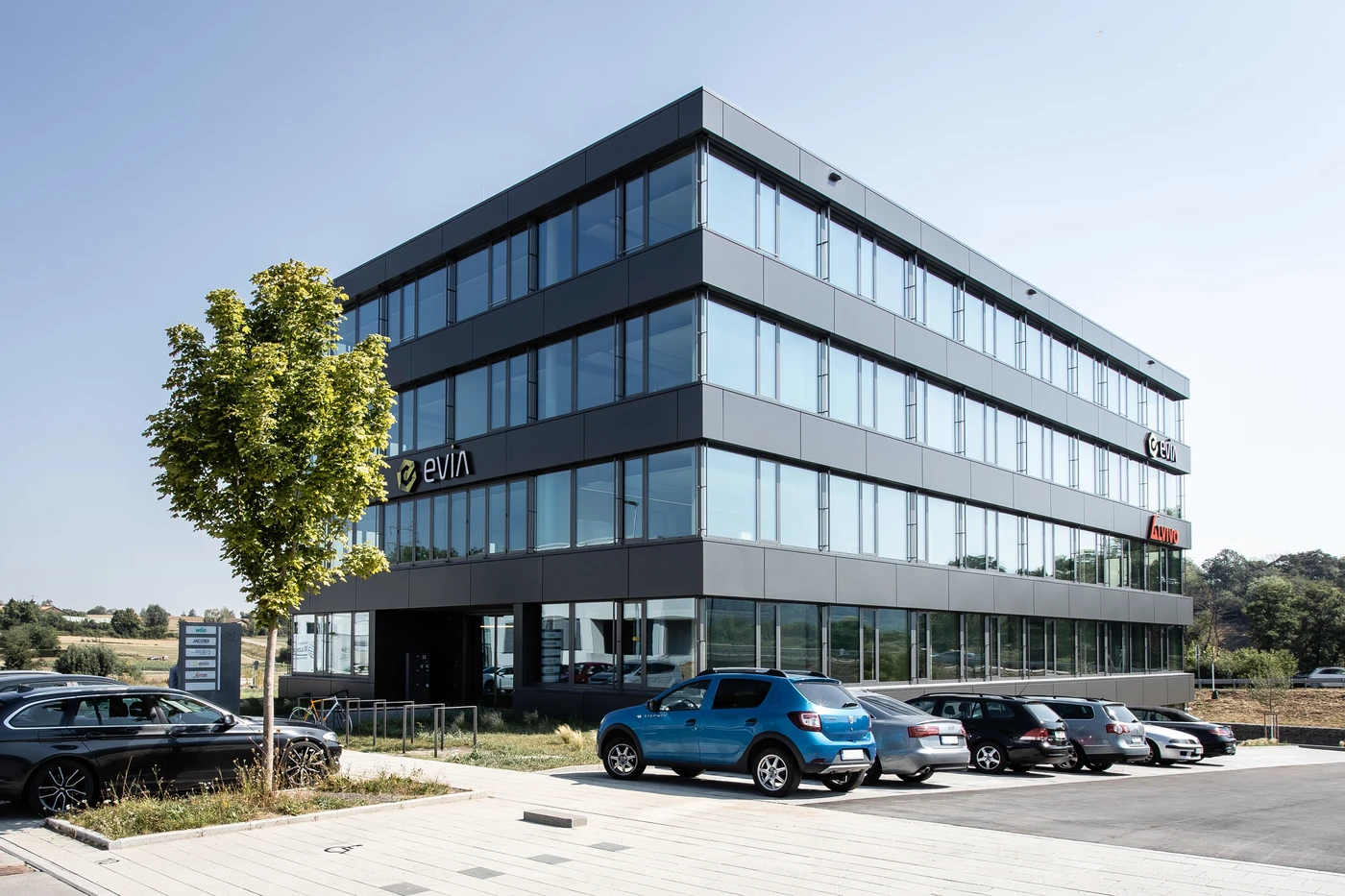 Ein modernes Bürogebäude aus Glas – entworfen unter Mitwirkung von SCHATZ Architektur – steht da, davor sind mehrere Autos geparkt, daneben steht ein kleiner Baum, und über ihm erstreckt sich ein klarer Himmel. An der Fassade sind die Namen „evin“ und „Acom“ zu sehen, was auf eine erfolgreiche Projektentwicklung über alle Leistungsphasen hinweg hinweist.