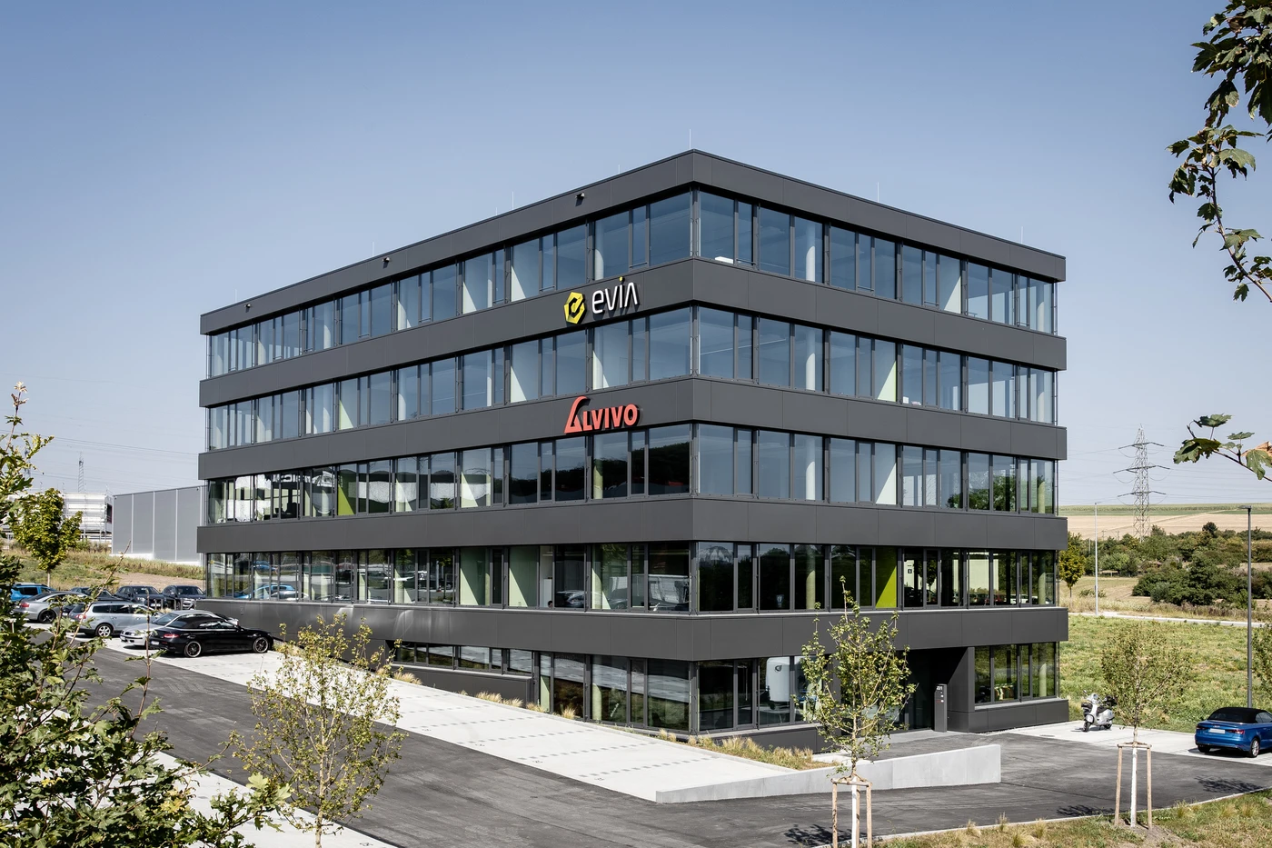 Generalplanung Ein modernes, vierstöckiges Bürogebäude mit großen Glasfenstern, an denen die Logos von eViva und Avivo zu sehen sind. Das von SCHATZ Architektur als innovativer Entwurf für Schorndorf entworfene Gebäude ist umgeben von jungen Bäumen, einem Parkplatz und offenen Rasenflächen unter freiem Himmel.