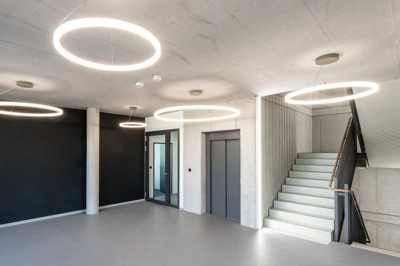 Projekte Moderne Lobby mit runden Deckenleuchten, einer schwarzen Akzentwand, Glastüren, einem Aufzug und einer Treppe mit dunklem Geländer, die nach oben führt. Der Raum spiegelt die Prinzipien der Generalplanung und einer durchdachten Projektentwicklung wider und zeichnet sich durch ein minimalistisches Design mit Betonböden und neutralen Farben aus.