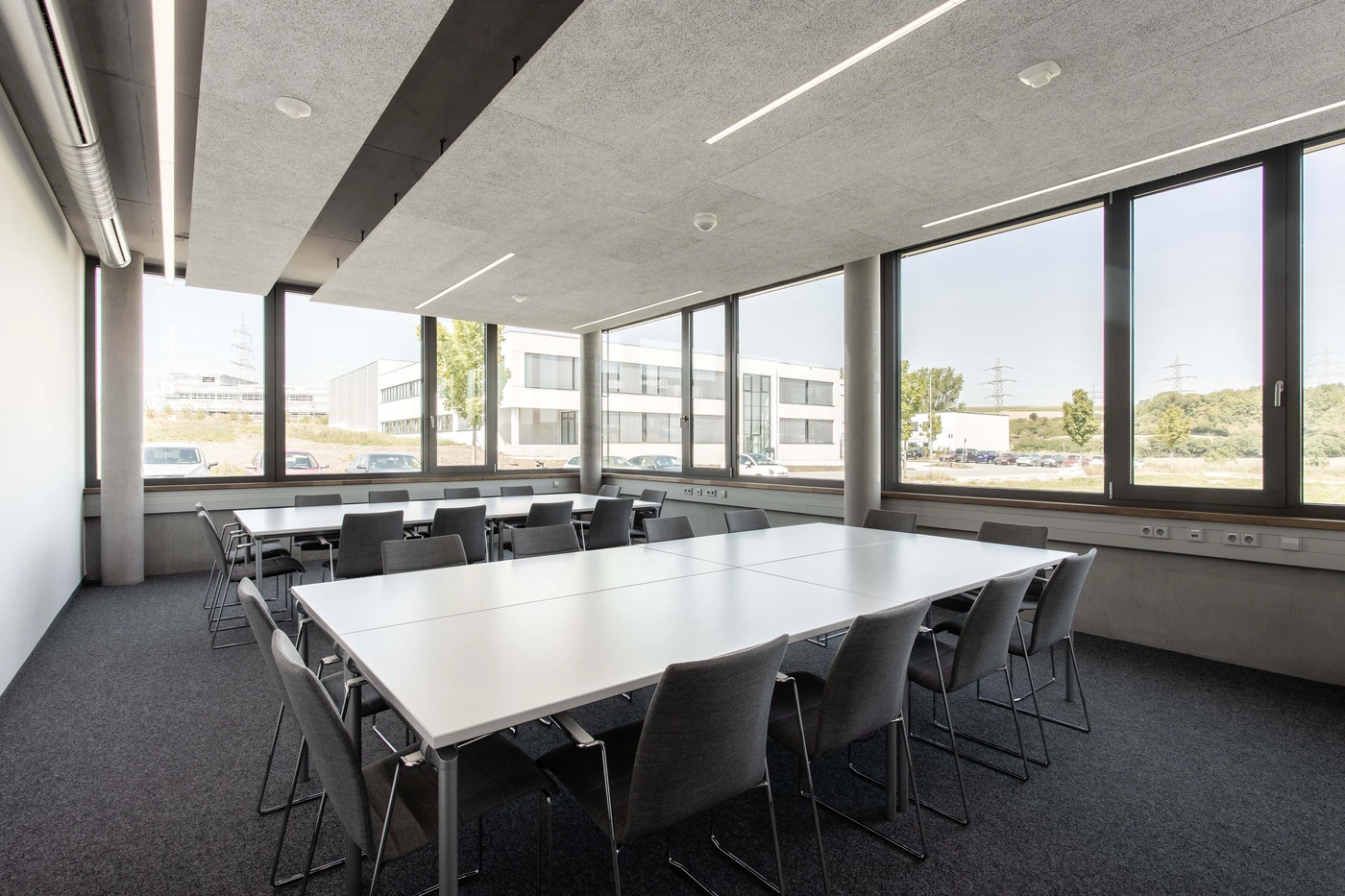 Ein moderner, von SCHATZ Architektur entworfener Konferenzraum mit großen Fenstern, zwei aneinandergereihten weißen Tischen und grauen Stühlen, die diese umgeben. Der Raum ist leer und hell, und von innen sind Bürogebäude zu sehen – ein Beispiel für durchdachten Entwurf und sorgfältige Projektentwicklung.