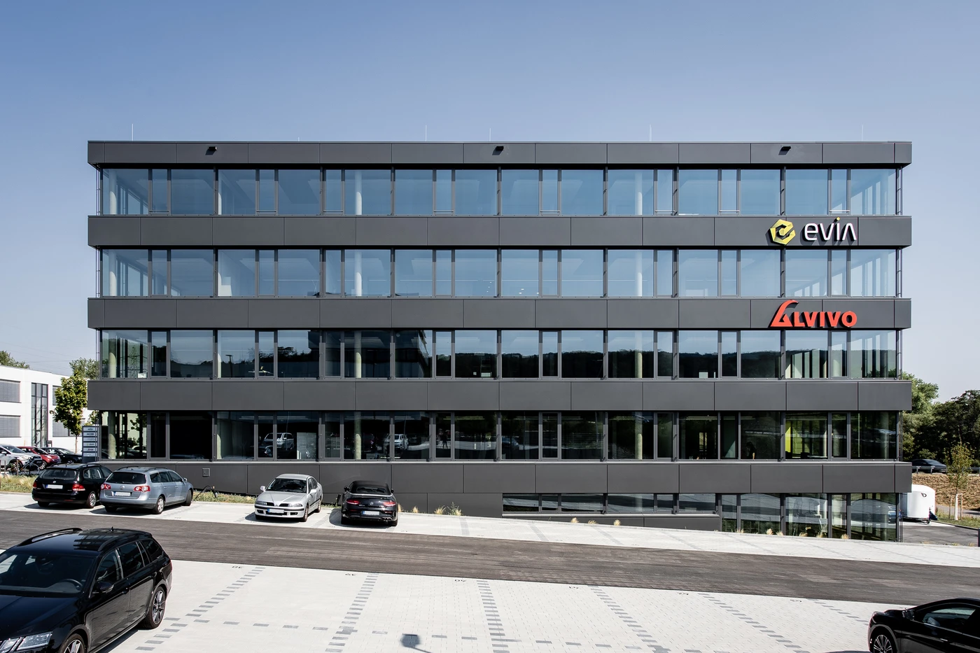 Projekte Ein modernes, graues Bürogebäude mit großen Fenstern, an denen die Logos von evia und Avivo zu sehen sind, präsentiert den raffinierten Entwurf von SCHATZ Architektur. An einem sonnigen Tag säumen mehrere geparkte Autos die Fassade und unterstreichen die Liebe zum Detail, die sich durch alle Leistungsphasen zieht.