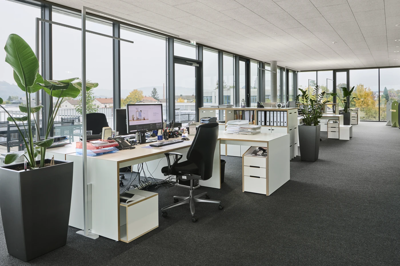 Ein modernes Großraumbüro mit großen Fenstern, mehreren Schreibtischen mit Computern, Bürostühlen, Unterlagen und Topfpflanzen. Der Raum ist hell, übersichtlich und bietet einen Blick auf die Stadtlandschaft mit Bäumen und Gebäuden – ein idealer Rahmen für Generalplanung oder Projektentwicklung im pulsierenden Schorndorf.
