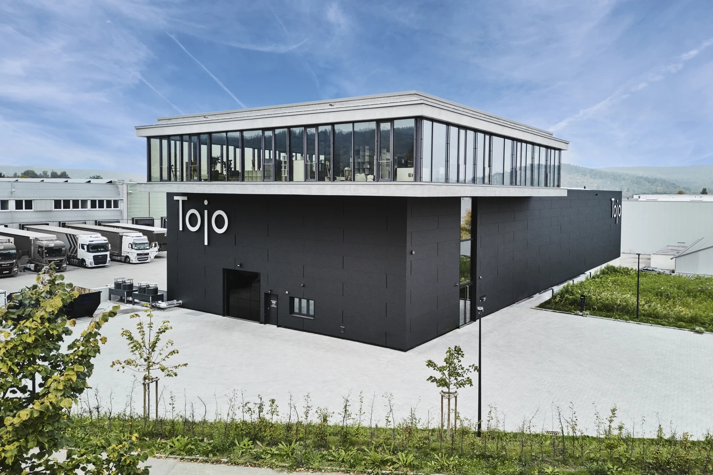 Modernes schwarzes Gebäude mit großen Fenstern im Obergeschoss, das den Namen „Tojo“ trägt. Es befindet sich in Schorndorf und ist umgeben von gepflasterten Gehwegen, Grünflächen und mehreren in der Nähe geparkten Lkw unter einem blauen Himmel mit leichten Wolken – ein hervorragendes Aushängeschild für Generalplanung über alle Leistungsphasen hinweg.